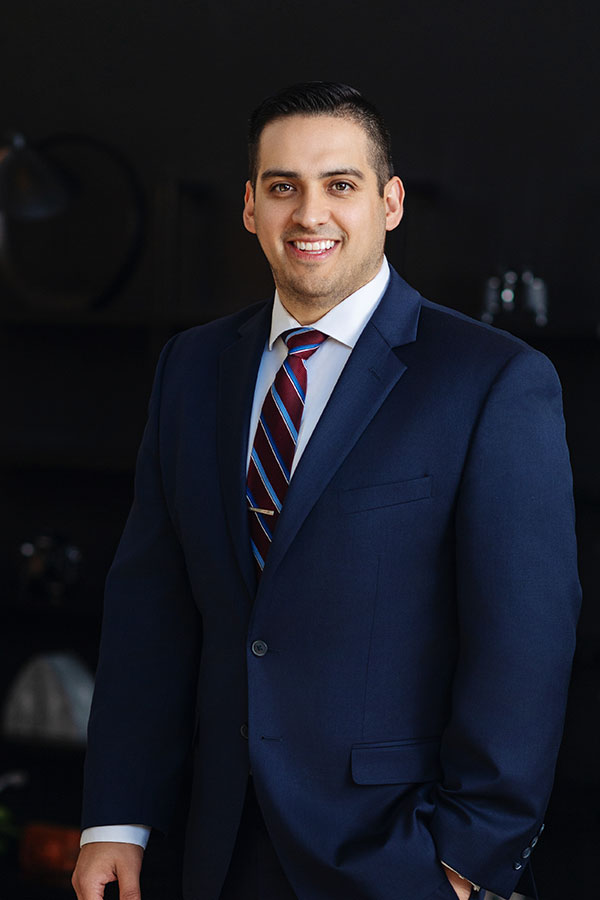 A professional waist-up portrait of attorney Alonzo Provencio. He is smiling and wearing a navy blue suit with a striped burgundy and blue tie, standing against a dark office background