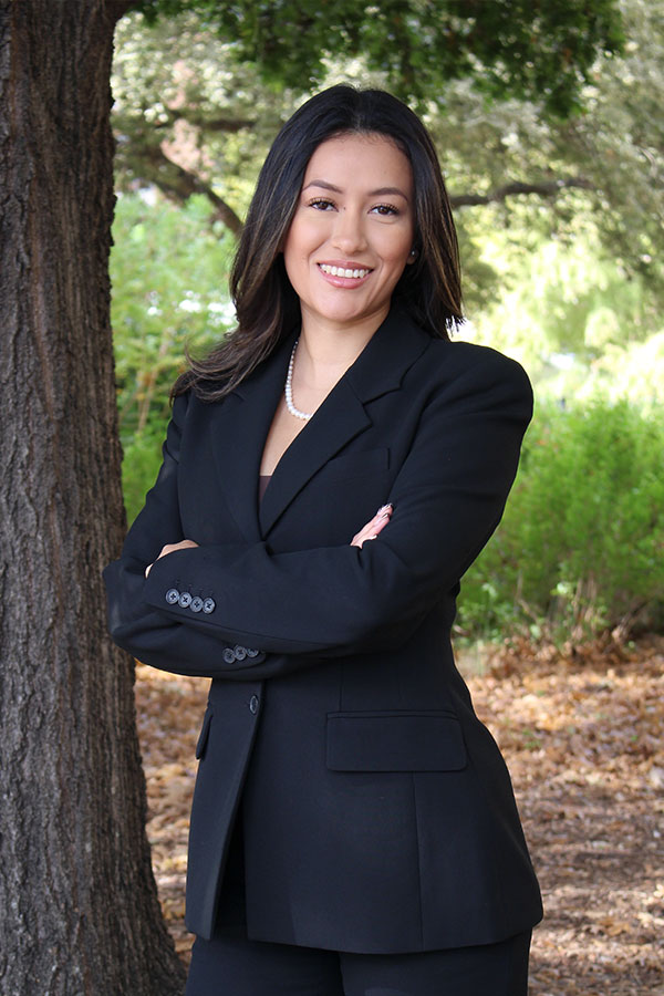 A professional waist-up portrait of attorney Ana Laura Rojas. She is smiling with her arms crossed, wearing a black blazer and pearl necklace, standing outdoors next to a large tree