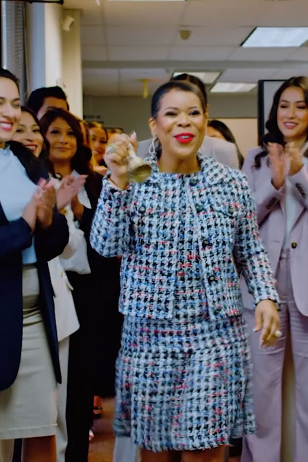 Erika Salter wearing a blue and white patterned suit, smiling and ringing a handheld bell while walking through her office as her staff claps and cheers in the background