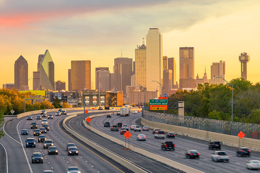 A busy multi-lane highway with traffic flowing toward the downtown Dallas skyline, including Reunion Tower, under a golden sunset sky